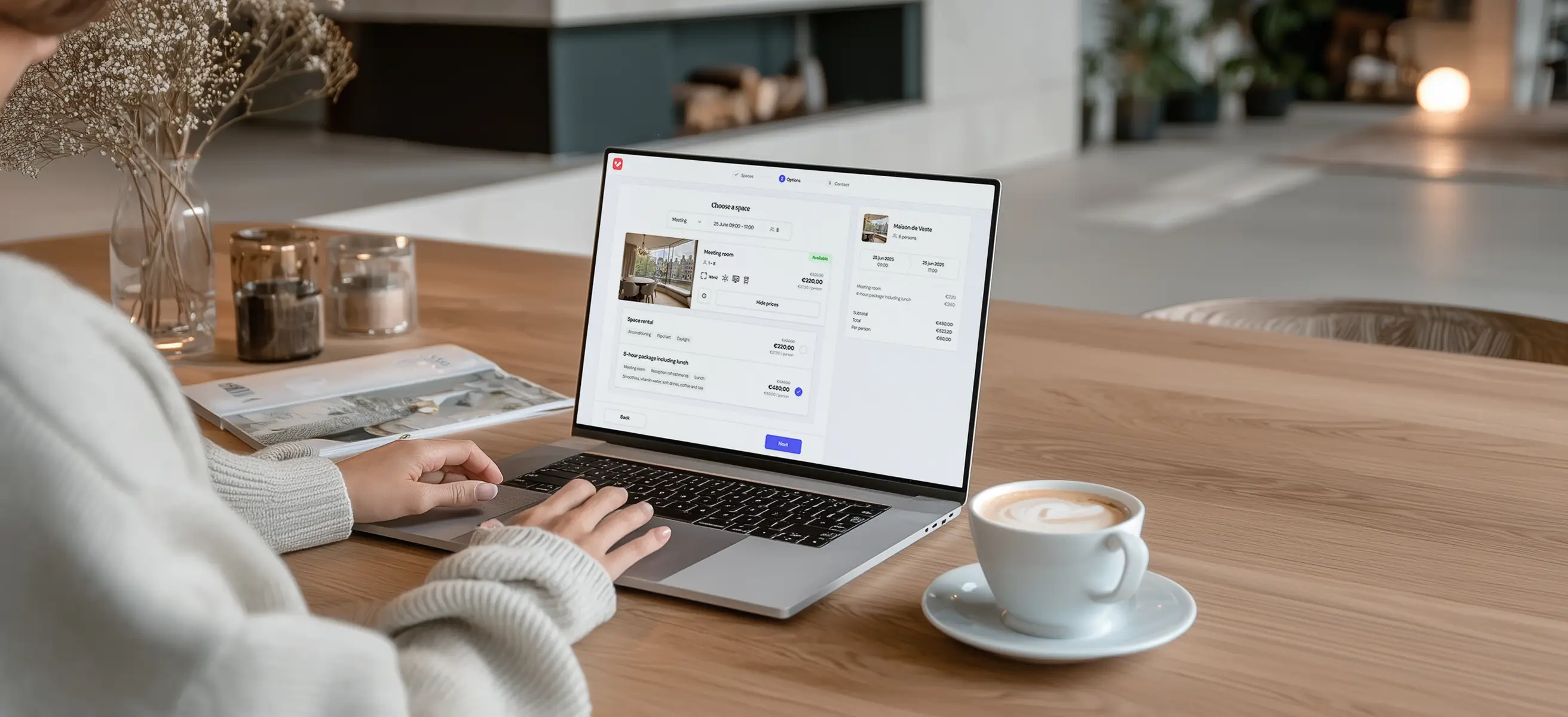 A venue host sits at home at the dining table with a laptop and a cup of coffee. The VenueSuite software is displayed on the laptop screen.