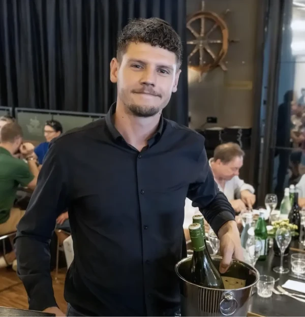 Man met een zwart overhemd en wijnflessen in zijn hand die in een druk restaurant staat